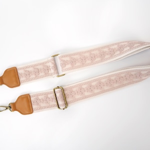 May include: A light pink adjustable bag strap featuring a textured floral lace pattern. The strap includes tan leather end tabs and antique brass-toned metal hardware, including two swivel lobster clasps and a sliding buckle for length adjustment.