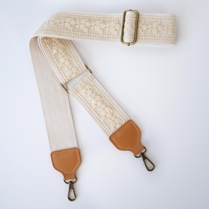 Puede incluir: Una correa de bolso ajustable de color crema con una franja central de intrincado encaje floral. La correa incluye extremos de piel sintética de color tostado y herrajes metálicos en tono latón antiguo, incluyendo una hebilla deslizante y dos mosquetones.