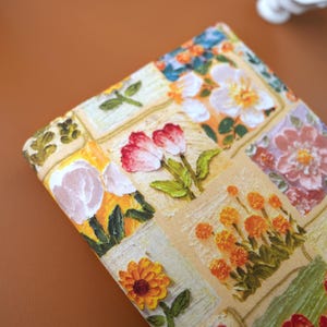 Puede incluir: Primer plano de una tela estampada con flores. El dise&ntilde;o presenta un mosaico de cuadrados con varias flores en tonos rojos, naranjas, amarillos y blancos. Tambi&eacute;n hay hojas y tallos verdes, sobre un fondo beige.