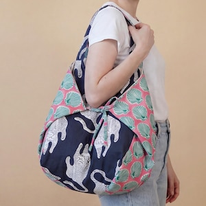 Könnte beinhalten: Eine Schultertasche im Hobo-Stil aus dunkelblauem Stoff mit weißem Tigermuster und kontrastierenden Seitenteilen aus rosa Stoff, der mit einem sich wiederholenden grünen Muscheldruck verziert ist. Die Tasche hat einen Bindebandverschluss.