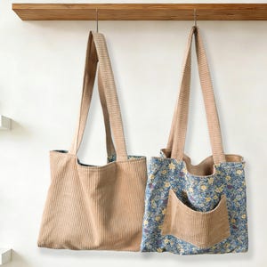 May include: Two corduroy tote bags hanging against a white wall. One bag is solid tan, the other has a blue floral pattern with a tan pocket. Both have long tan straps and are suspended from a wooden shelf.