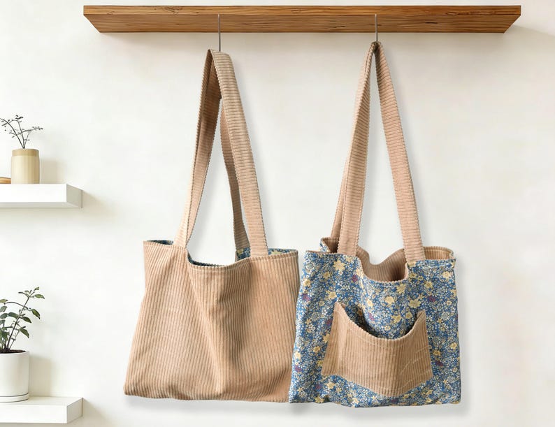 May include: Two corduroy tote bags hanging against a white wall. One bag is solid tan, the other has a blue floral pattern with a tan pocket. Both have long tan straps and are suspended from a wooden shelf.