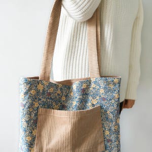 May include: A floral tote bag with a blue background and yellow flowers. The bag has a tan corduroy pocket and matching corduroy straps. The bag is held by a person wearing a white sweater.