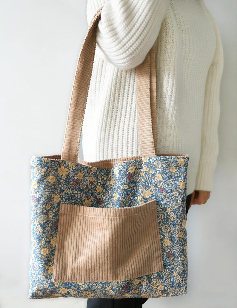 May include: A floral tote bag with a blue background and yellow flowers. The bag has a tan corduroy pocket and matching corduroy straps. The bag is held by a person wearing a white sweater.