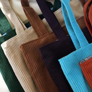 May include: A collection of corduroy tote bags in various colors, including green, beige, brown, navy, turquoise, and rust. Each bag has matching corduroy straps.