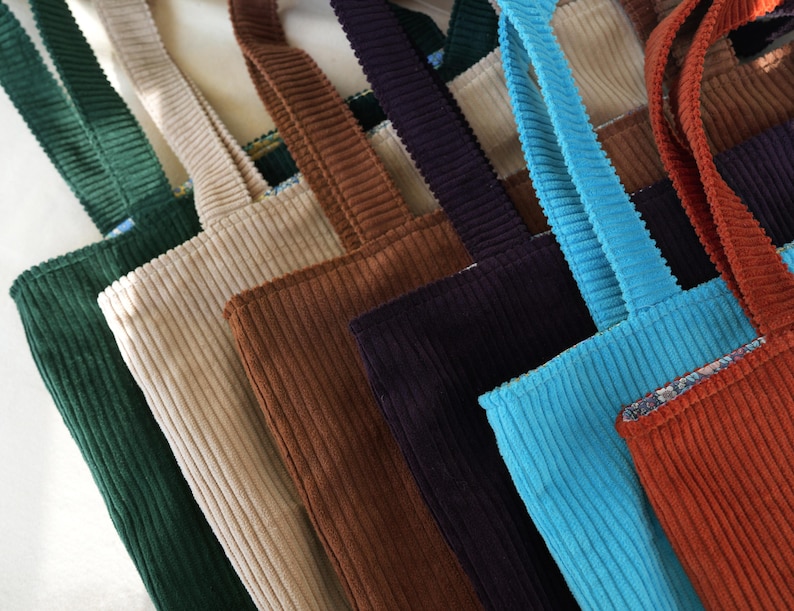 May include: A collection of corduroy tote bags in various colors, including green, beige, brown, navy, turquoise, and rust. Each bag has matching corduroy straps.