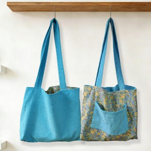 May include: Two turquoise corduroy tote bags hanging against a white wall. One bag is solid turquoise, the other has a floral pattern with a turquoise pocket. Both have long, matching corduroy straps.