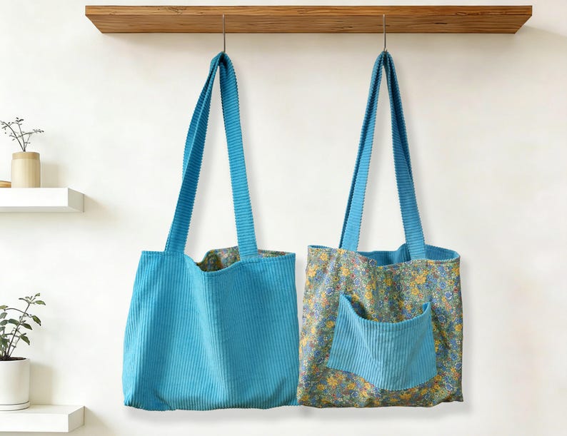 May include: Two turquoise corduroy tote bags hanging against a white wall. One bag is solid turquoise, the other has a floral pattern with a turquoise pocket. Both have long, matching corduroy straps.