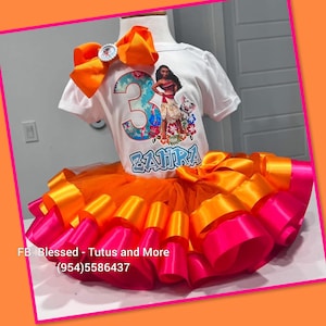 Op de afbeelding: Een wit T-shirt met korte mouwen voorzien van een afbeelding van een personage, het cijfer 3 en de naam ZAHRA, gecombineerd met een oranje en roze gelaagde tule tutu-rok. Een grote oranje haarstrik is bevestigd aan de halslijn van het shirt.