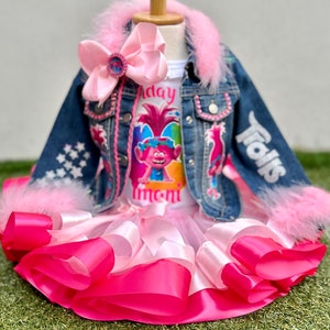 May include: A pink and white tulle tutu skirt with a pink satin ribbon trim. A denim jacket with pink fur trim and the word "Trolls" on the back. A white t-shirt with a Trolls character and the text "Birthday Girl".