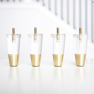 May include: Four clear acrylic furniture legs with gold metal bases. The legs are tapered and have a screw thread at the top.
