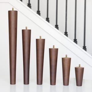 May include: A set of six wooden furniture legs in varying heights. The legs are a dark brown color with a brass-colored metal accent at the top. The legs are arranged in descending order of height, showcasing their tapered design.