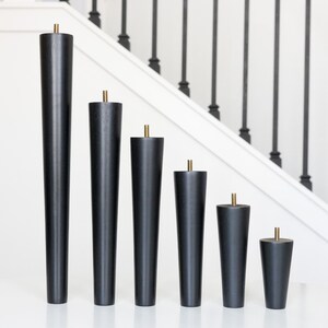 May include: Six black wooden furniture legs of varying heights, each with a brass-colored metal accent at the top. The legs are arranged in a descending order, showcasing their different sizes. The background features a white staircase with black railings.