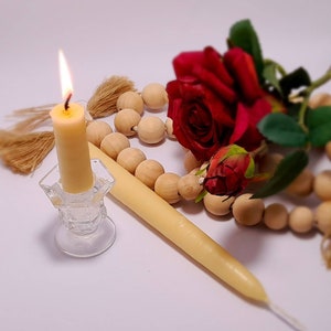 Pu&ograve; includere: Una candela color crema accesa in un portacandele di vetro trasparente, una seconda candela spenta, perline di legno e una rosa rossa. La fiamma della candela &egrave; gialla. Gli oggetti sono disposti su una superficie bianca.