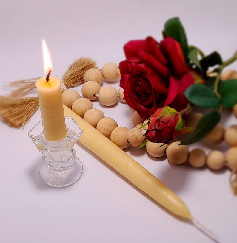 Pu&ograve; includere: Una candela color crema accesa in un portacandele di vetro trasparente, una seconda candela spenta, perline di legno e una rosa rossa. La fiamma della candela &egrave; gialla. Gli oggetti sono disposti su una superficie bianca.