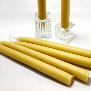 Pu&ograve; includere: Una collezione di candele affusolate gialle, alcune distese e altre in portacandele quadrati di vetro trasparente. Le candele hanno una forma cilindrica liscia e sono di un caldo colore giallo.