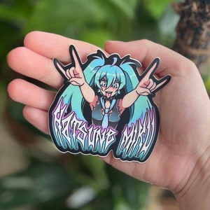 May include: A sticker featuring a cartoon character with teal hair, wearing a black and blue outfit, and making a rock-on hand gesture. The sticker has the text "Hatsune Miku" in a stylized font with a black outline.