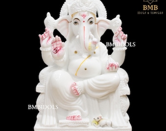 May include: White marble statue of Ganesha, the Hindu god of wisdom and prosperity, sitting on a throne with a mouse at his feet. The statue is decorated with gold and red paint.