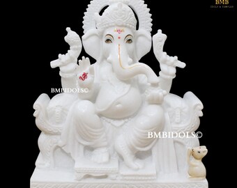 May include: White marble statue of Ganesha, the Hindu god of wisdom and prosperity, sitting on a throne with a mouse at his feet. The statue is intricately carved with details and features a golden halo and a red Om symbol on his hand.