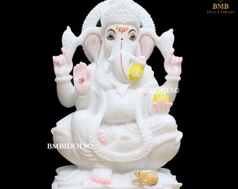 May include: White marble statue of Ganesha, the Hindu god of wisdom and prosperity, sitting in a meditative pose with his trunk curled and holding a sweet in his hand. The statue is decorated with pink and yellow accents and features a small frog at the base.
