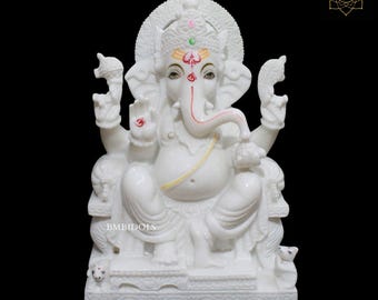 May include: White marble statue of Ganesha, the Hindu god of wisdom and prosperity, sitting on a throne with his trunk curled up. He is adorned with colorful decorations and a yellow sash.