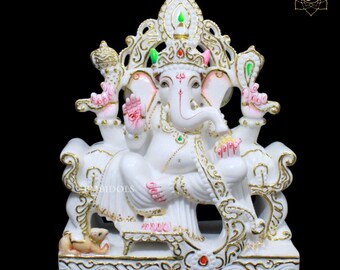 May include: White marble statue of Ganesha, the Hindu god of wisdom and prosperity, sitting on a throne with a crown and ornate details. The statue is decorated with gold accents and pink and green paint.