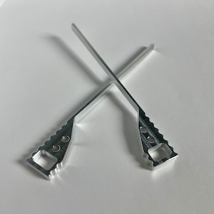 Może przedstawiać: Dwa srebrne metalowe narzędzia barowe skrzyżowane na białym tle. Każde narzędzie ma prostokątny uchwyt z kwadratowym otworem i ząbkowaną krawędzią. Pręty są cienkie i proste.