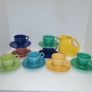 Może przedstawiać: Zestaw ceramicznej zastawy stołowej składający się z sześciu kolorowych filiżanek ze spodkami w odcieniach jasnej zieleni, błękitu, granatu, żółci i różu oraz jednego żółtego dzbanka. Każdy element ma gładkie wykończenie i okrągłe ucho.
