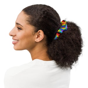 May include: A woman with dark curly hair wearing a white shirt and a rainbow patterned hair tie.
