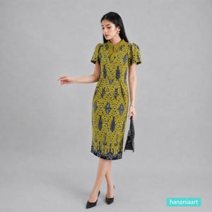 Puede incluir: Un vestido hasta la rodilla en color verde oliva con un patrón ikat geométrico azul oscuro, mangas cortas abullonadas, cuello mao y cintura entallada. La prenda se combina con un pequeño bolso negro, mostrando una silueta estructurada.