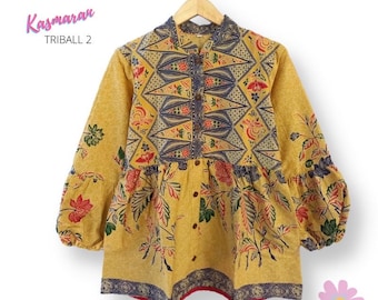 May include: A yellow, blue, and red patterned blouse with a button-up front and long sleeves. The blouse has a gathered waist and a floral print. The text "Kasmaran TRIBALL 2" is visible in the top left corner of the image.