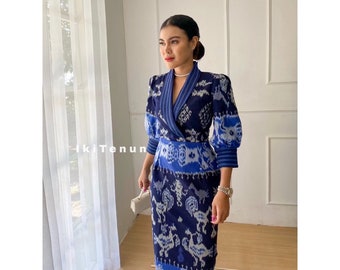 Può includere: Un abito a fantasia blu e bianca con scollo a V e maniche lunghe. L'abito ha uno stile avvolgente e una silhouette aderente. Il tessuto è un ikat tessuto, con un motivo geometrico. L'abito è mostrato su una modella con una pochette bianca.