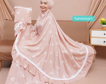 May include: A light pink prayer rug with white floral embroidery and a white lace trim. The rug is folded and has a matching carrying bag.