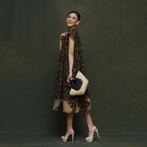Pode incluir: Uma mulher vestindo um vestido preto e marrom estampado com uma capa combinando. Ela está segurando uma bolsa de palha com uma alça preta. Ela está usando saltos plataforma. O fundo é verde escuro.
