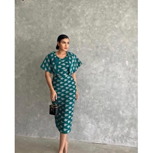 Puede incluir: Un vestido azul verdoso con un patrón blanco y una chaqueta a juego azul verdoso con ribetes blancos. El vestido y la chaqueta están hechos de tejido tejido. La modelo lleva un pequeño bolso de mano negro.