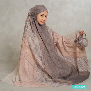 May include: A prayer garment featuring a hood and a flowing silhouette in shades of beige, taupe, and brown. The fabric displays a repeating pattern of chains, plaid, and subtle textures. A matching small fabric bag is held in the right hand.