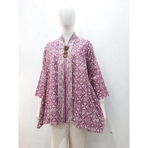 May include: A purple and white patterned blouse with a decorative button on the front. The blouse has a loose fit and a slightly asymmetrical hemline.