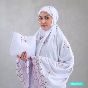May include: A white prayer garment featuring a matching head covering and a rectangular pouch. The fabric is decorated with embroidered pink flowers and brown vines, with scalloped edges finished in pink thread along the sleeves and hem.