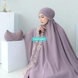 May include: A lilac prayer garment with intricate lace detailing, alongside a matching handbag with a gold chain strap. The image includes the text "hananiaart". The garment is draped over a person.