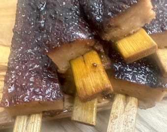 May include: Close-up of several pieces of food on wooden sticks. The food has a dark, glossy exterior and a lighter interior. The wooden sticks are a light brown color, and the food is arranged on a wooden surface.