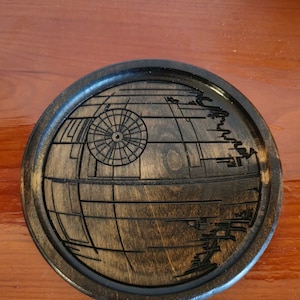 May include: A round wooden coaster with a dark brown stain. The coaster features a laser-etched design of the Death Star from Star Wars.