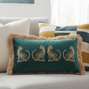 May include: A rectangular teal throw pillow with four embroidered cheetahs. The pillow has a tan fringe border and is placed on a light gray sofa. Other pillows with geometric patterns are in the background.