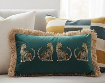 May include: A rectangular teal throw pillow with four embroidered cheetahs. The pillow has a tan fringe border and is placed on a light gray sofa. Other pillows with geometric patterns are in the background.