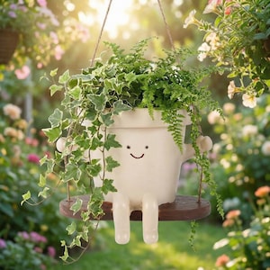 May include: A whimsical, off-white hanging planter with a smiling face, arms, and legs, holding a wooden swing. The planter is filled with green plants and trailing ivy. The background is a blurred garden scene with flowers.