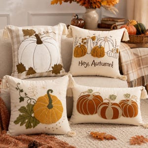 May include: Four decorative throw pillows featuring pumpkin designs. One has a large white pumpkin on a beige plaid background. Another says Hey, Autumn with embroidered pumpkins. Two others feature orange and brown pumpkin motifs with tassels.