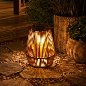 Può includere: Una lanterna da esterno cilindrica costruita con canne di rattan marrone verticali, dotata di una fonte di luce calda all'interno e di un pannello solare integrato nella maniglia superiore, che proietta ombre lineari sul terreno.