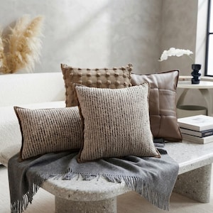 May include: A collection of four decorative throw pillows in neutral earth tones. One square pillow features a textured grid pattern, one square pillow has a smooth brown faux leather finish, and two rectangular pillows have a ribbed fabric texture.