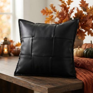May include: A square throw pillow featuring a black faux leather cover with a woven, basket-weave pattern. The surface consists of wide, interlocking strips of smooth, matte black material that create a grid design across the entire front face.