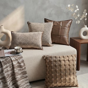 May include: A collection of four decorative throw pillows in neutral earth tones. One rectangular pillow features a ribbed texture, one square pillow has a woven grid pattern, one square pillow is smooth brown faux leather, and one has a tufted grid.