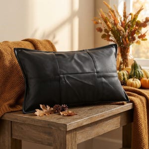 May include: A rectangular black leather pillow featuring a paneled construction with visible stitching. The pillow has a smooth, matte finish and is positioned on a wooden surface, accompanied by small brown leaves and pine cones.